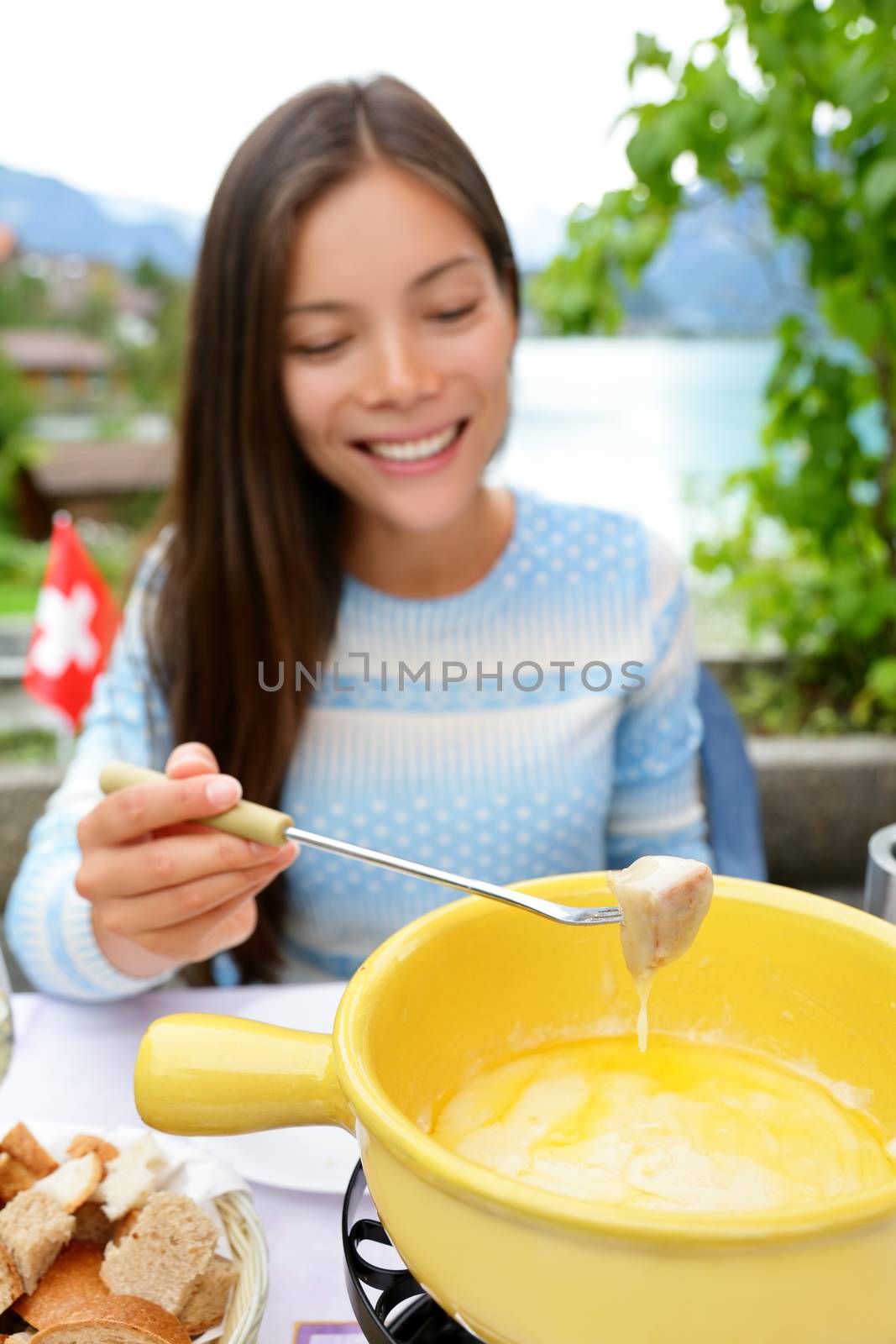 Cheese fondue - woman eating local Swiss food dipping bread in melted cheese. People eating traditional food from Switzerland having fun by lake in the Alps on travel in Europe.