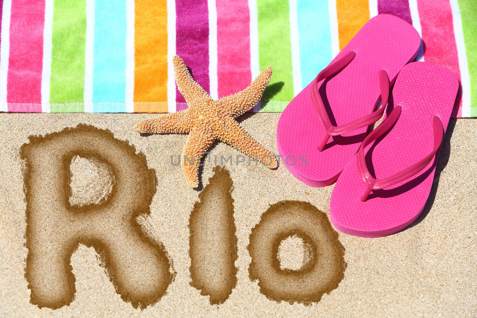 Rio. Overhead view ot the word RIO written on beach sand with a colorful striped towel, pink thongs and a starfish conceptual of a summer vacation and travel in Rio de Janiero, Brazil