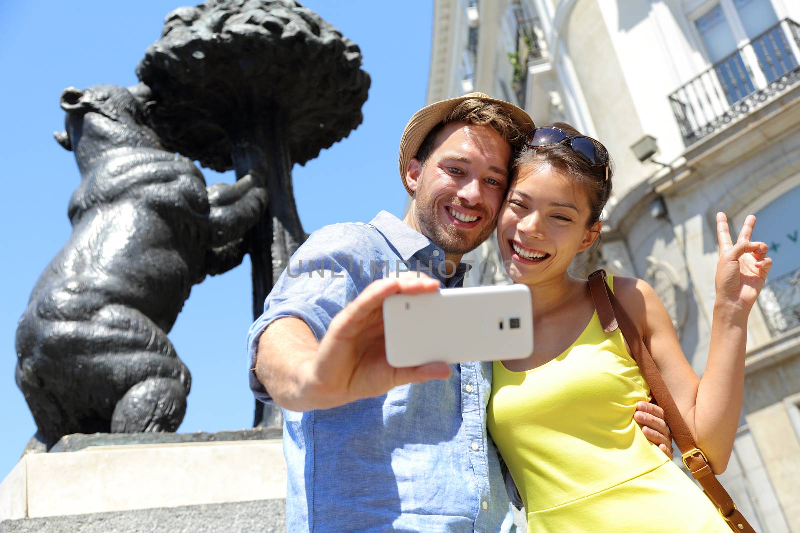 Tourists taking selfie photo pictures by famous bear statue Madrid on Puerta del Sol. Young couple using smartphone camera at tourist attraction Bear and the Madrono Tree, symbol of Madrid, Spain.