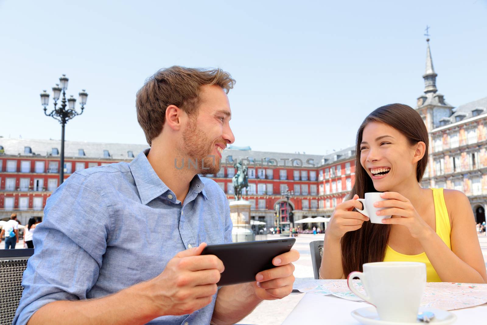 Madrid tourists at cafe drinking coffee having fun using tablet travel app on Plaza Mayor. Tourist couple sightseeing visiting tourism landmarks and attractions in Spain. Young woman and man
