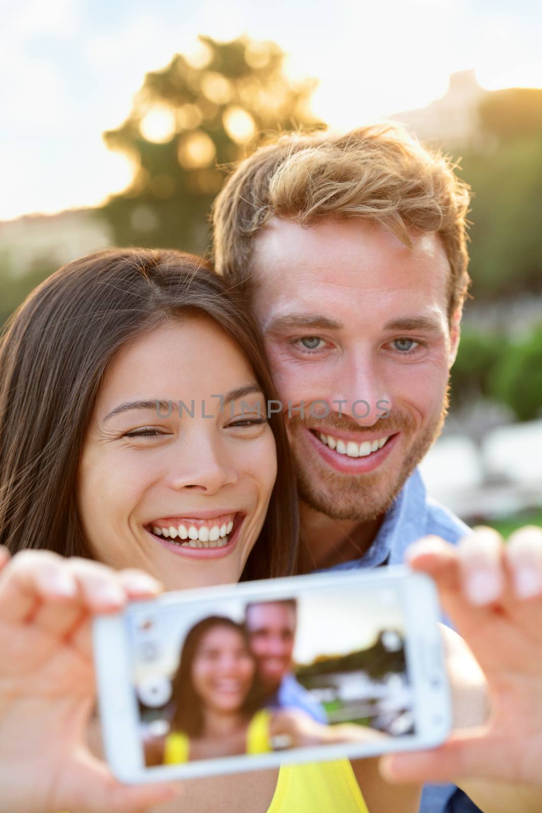 Couple in love taking selfie photo with smartphone. Romantic mixed race couple taking selfies pictures with smart phone camera having fun together outdoors in park. Asian girl, Caucasian guy.