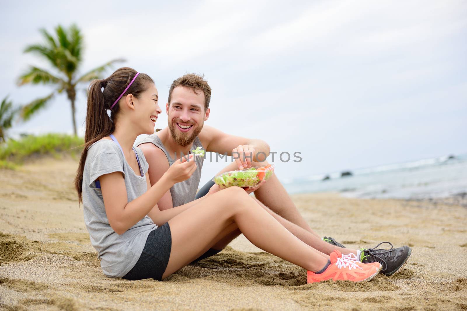 Salad - healthy fitness woman and man couple laughing eating food lunch sitting on beach after workout. Mixed race Asian Caucasian female model and male models in sportswear.