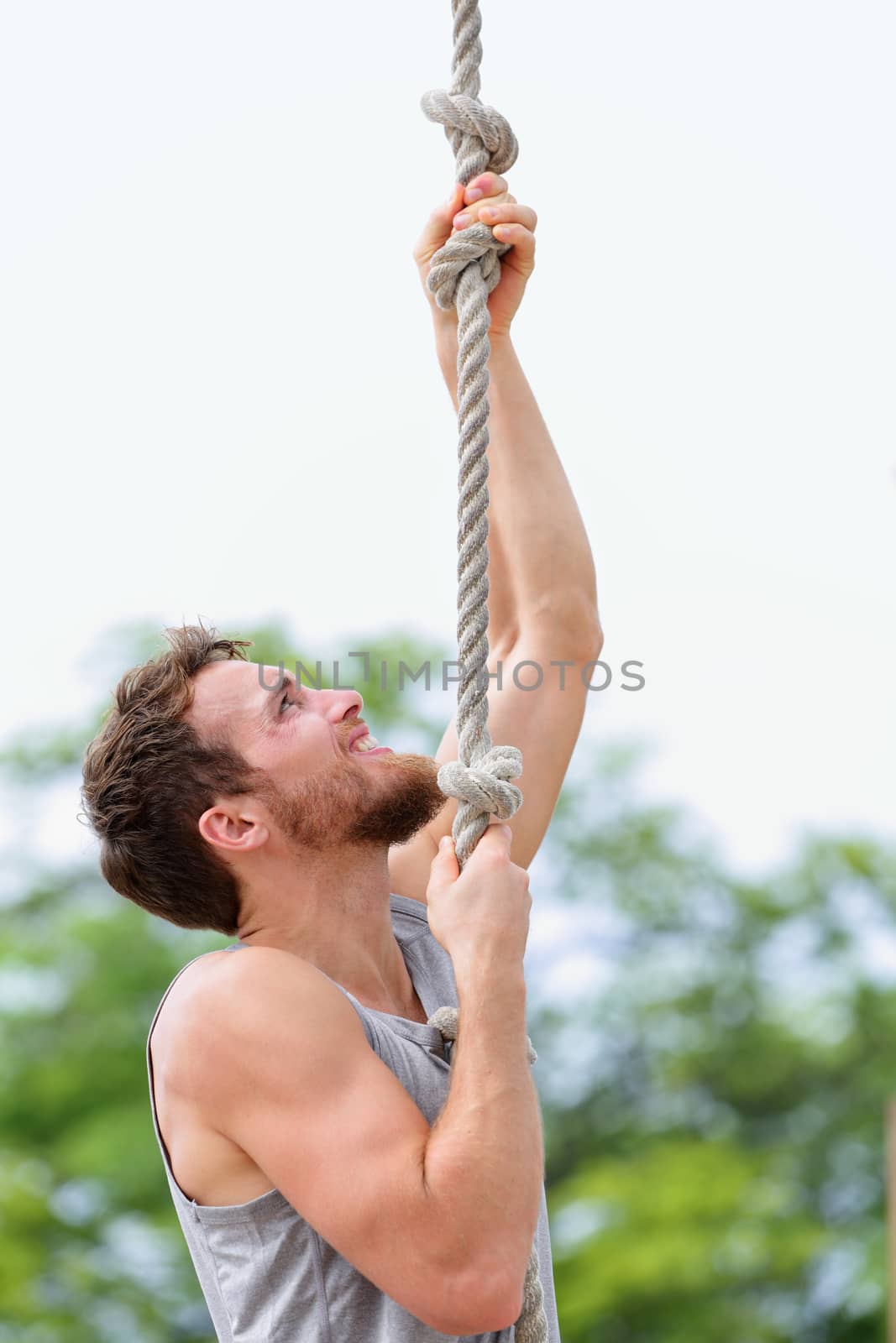 Crossfit man doing rope climb workout outside during summer. Handsome male athlete climbing up rope for strength training session.