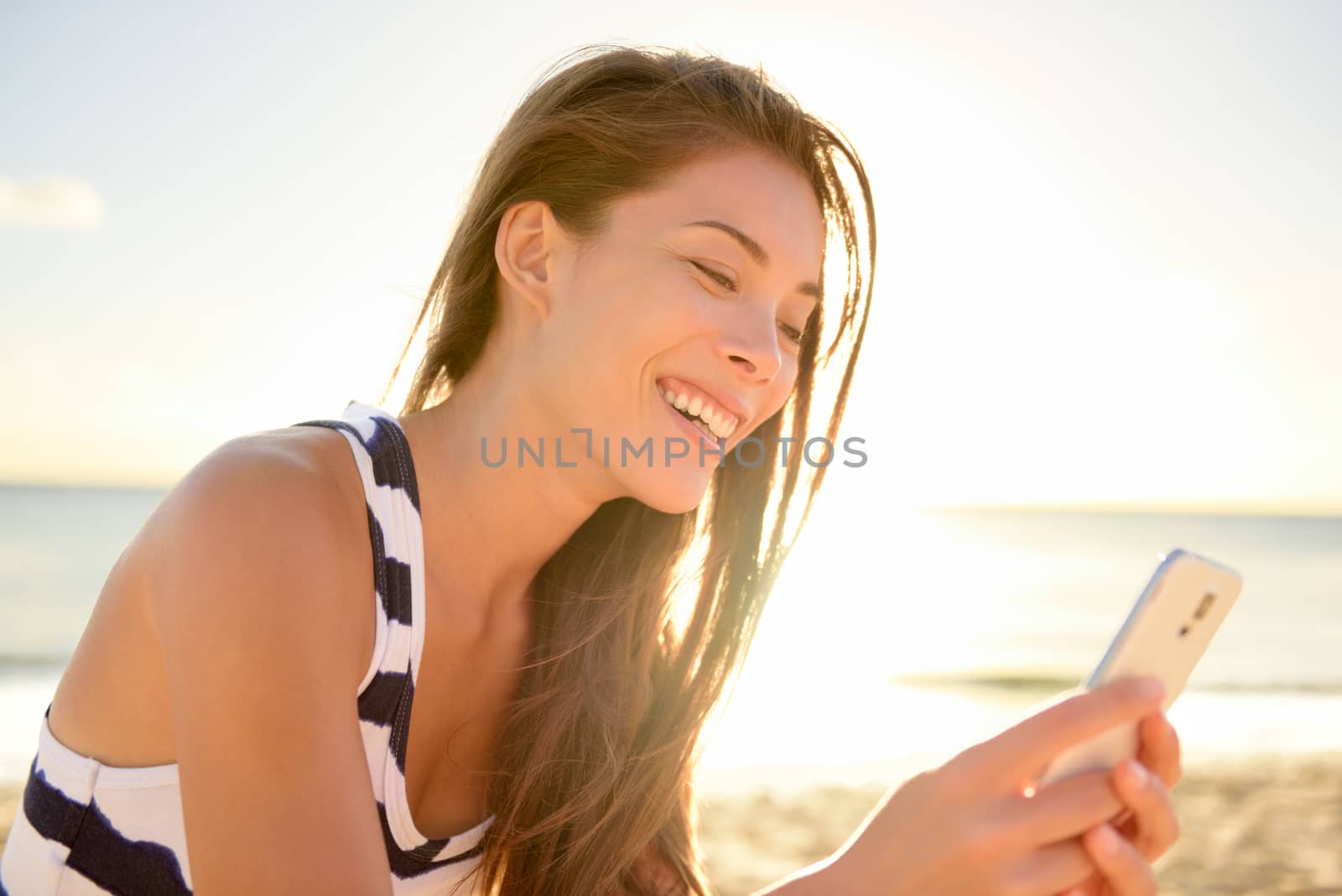 Mixed race pretty smiling young woman holding and looking at smart phone on hawaiian beach in sunset, enjoying summer vacation travel. Sea and horizon in background