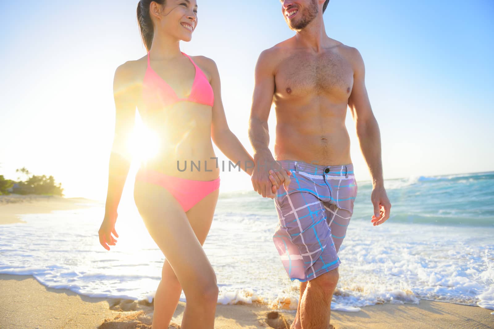 Mixed race couple walking holding hands happily smiling at beach sunset waterfront, enjoying summer honeymoon travel vacation. Hawaii