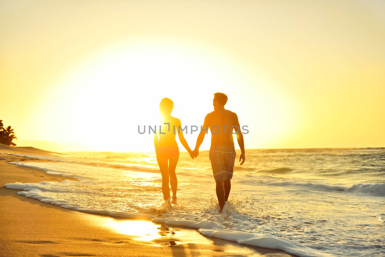 Honeymoon romantic couple in love holding hands walking on beautiful sunset at beach in waterfront. Lovers or newlywed married young couple by the sea enjoying relaxed vacation travel holiday. Hawaii.