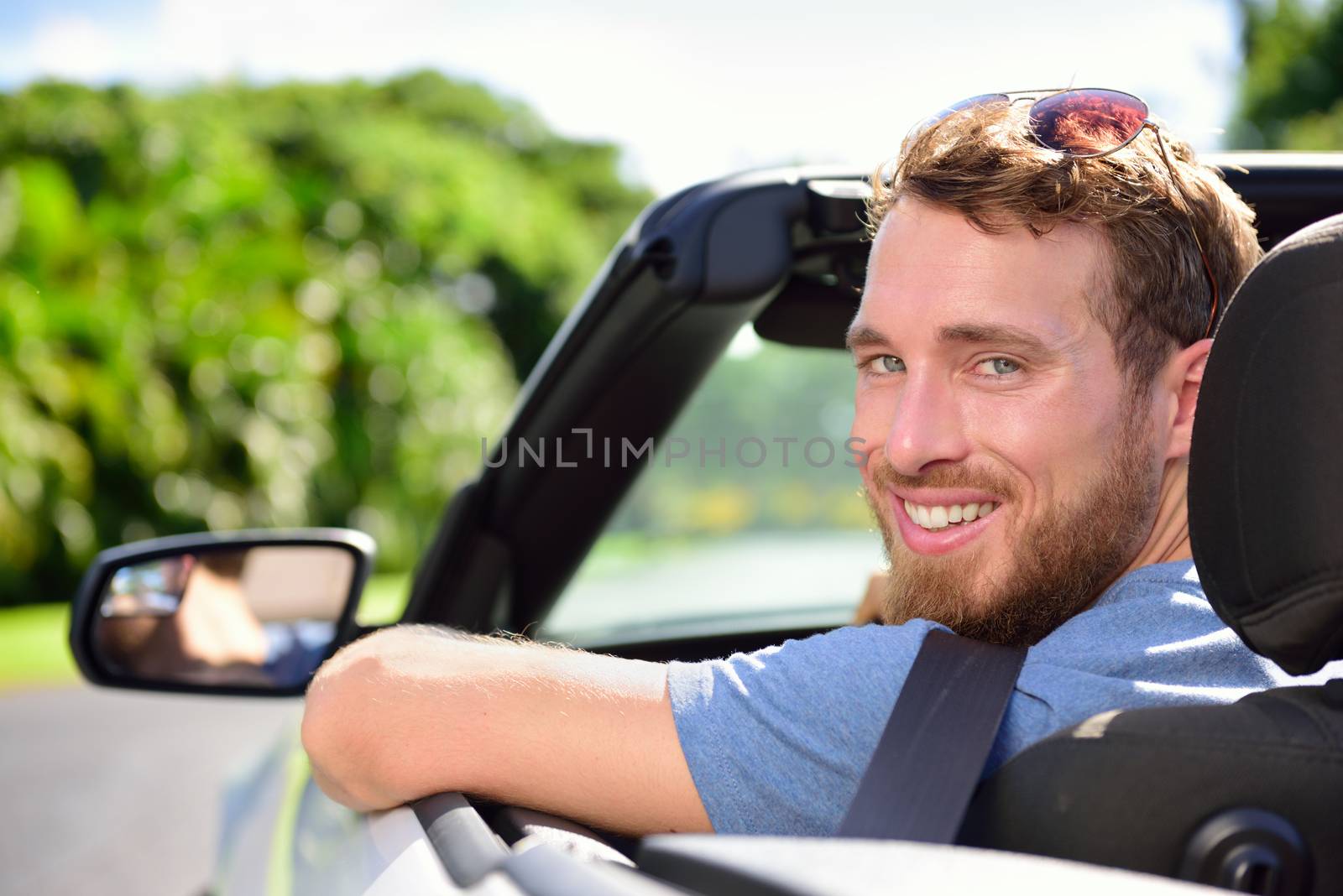 Car driver - young man wearing safety belt driving convertible on road trip in summer. Caucasian male looking at camera.