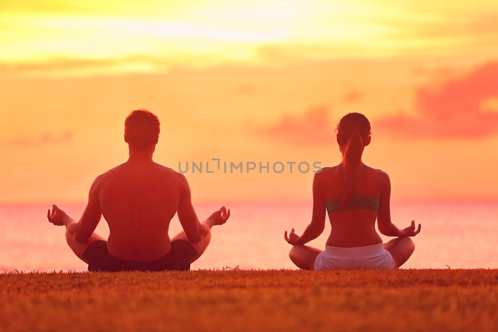 Meditation yoga couple meditating at serene beach sunset. Girl and man relaxing in lotus pose in calm zen moment in the ocean during yoga holiday class at resort retreat.