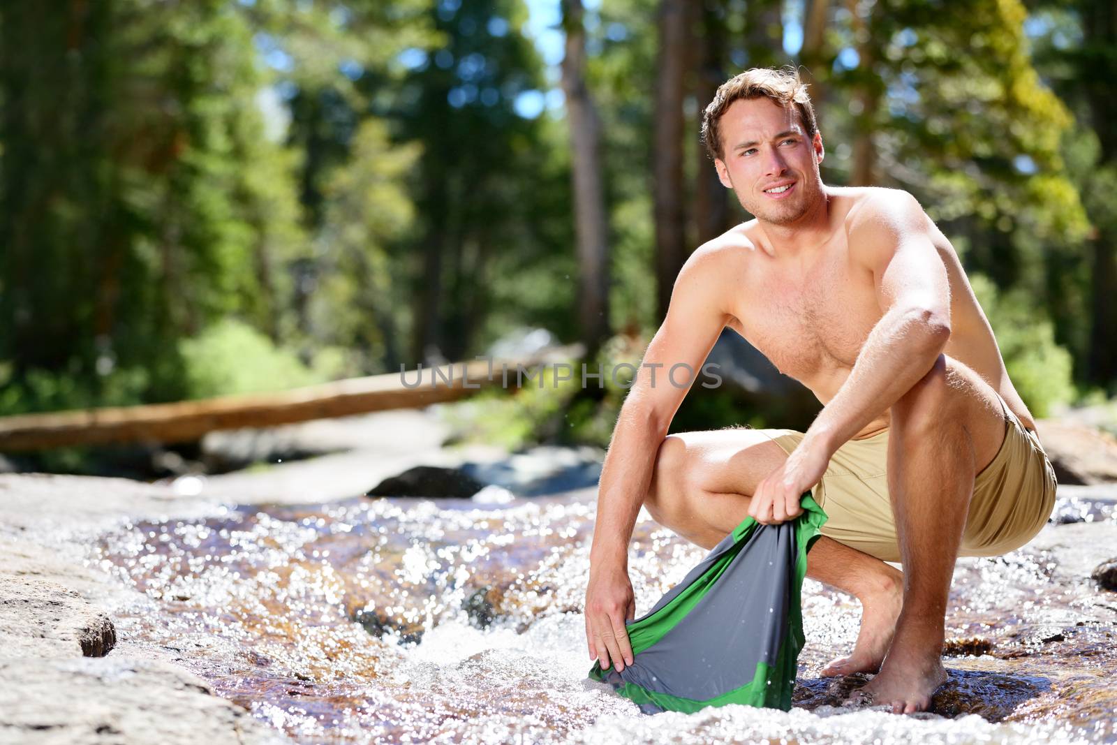 Camping hiker man washing clothes on trek in nature river. Hiking young male adult doing clothing wash chores in natural stream of water during an adventure trip outdoor.