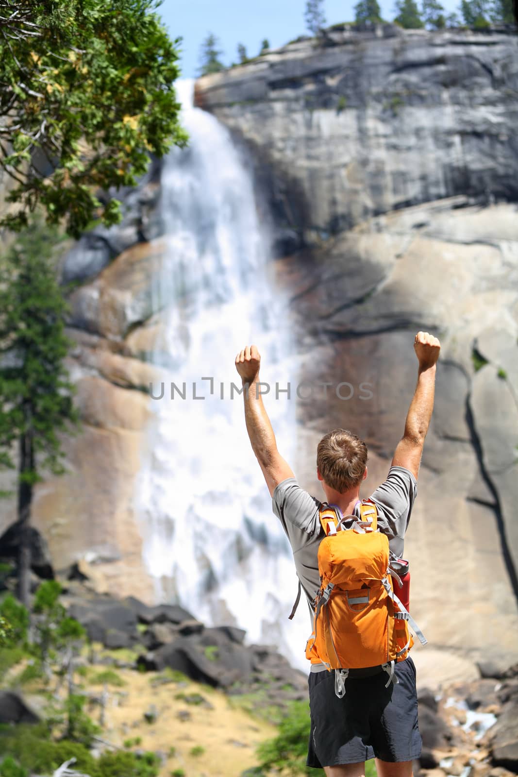 Happy hiking man cheering in success or freedom by waterfall in Yosemite national park. Winner hiker enjoying view of beautiful summer nature landscape, California, USA.