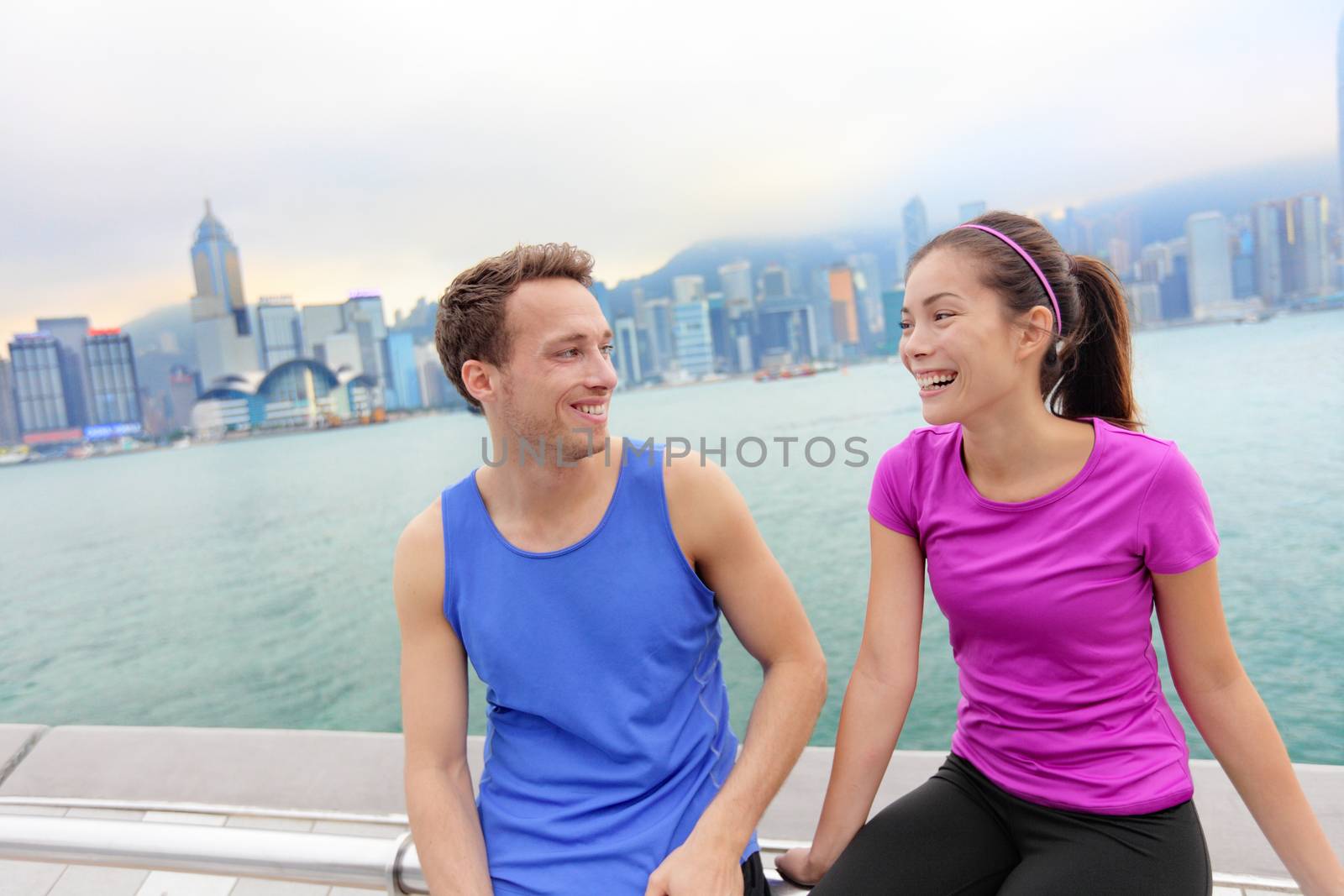 Runners relaxing after workout in Hong Kong city. Running caucasian and asian man and woman post run taking a break talking together on the Avenue of the Stars in Victoria harbor, HongKong Skyline.
