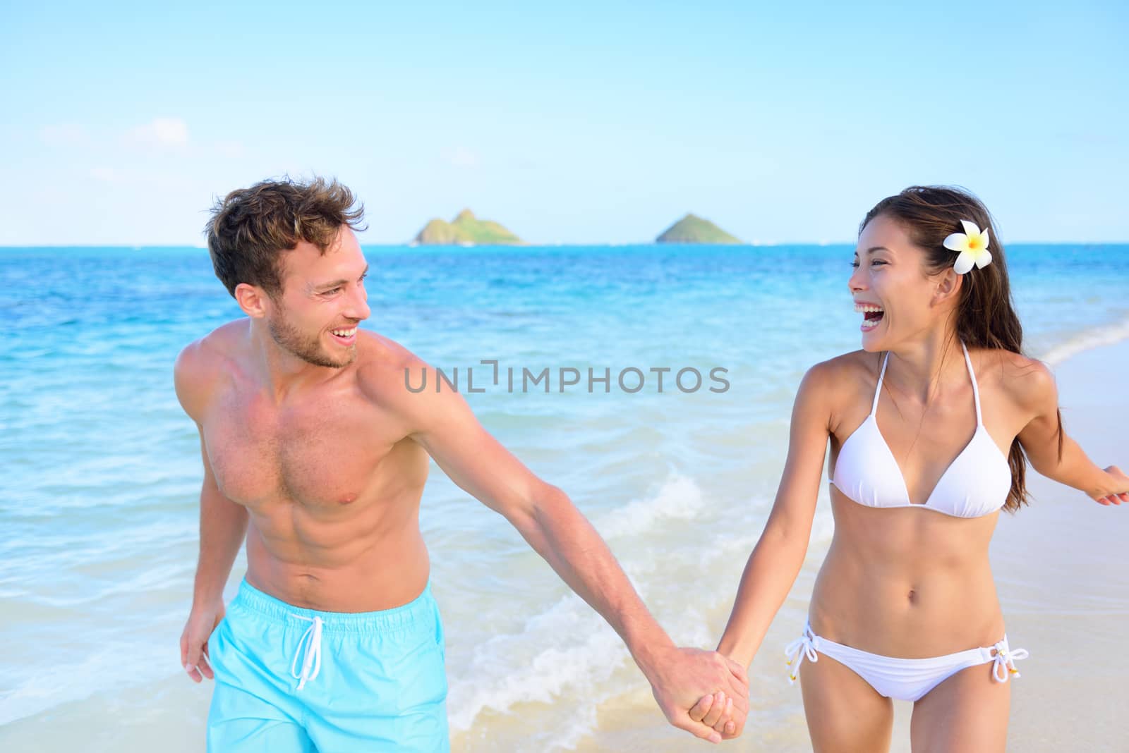 Fun on beach - couple in a happy relationship running together on a hawaiian beach during holidays travel vacation. Mixed race asian chinese woman and Caucasian man holding hands in trust in the sun.