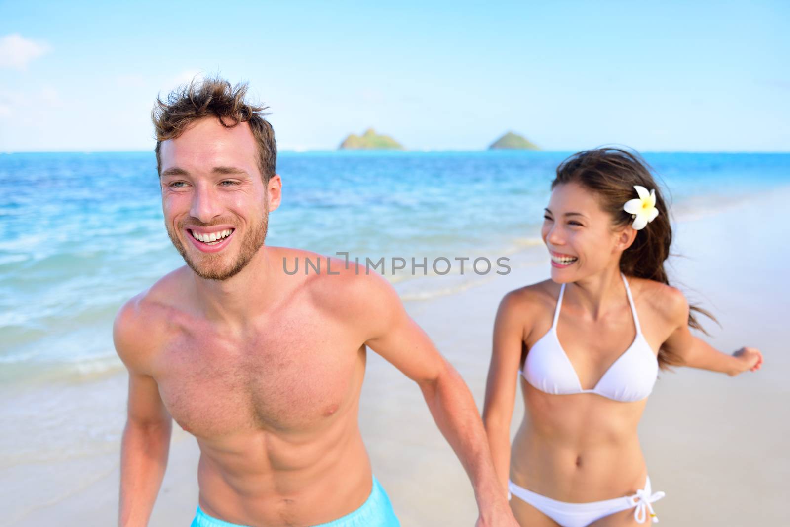 Happy couple having fun on beach vacation during summer holiday. Multiracial fit couple running together holding hands laughing in the sun. Young adults in shape carefree feeling good in their body.