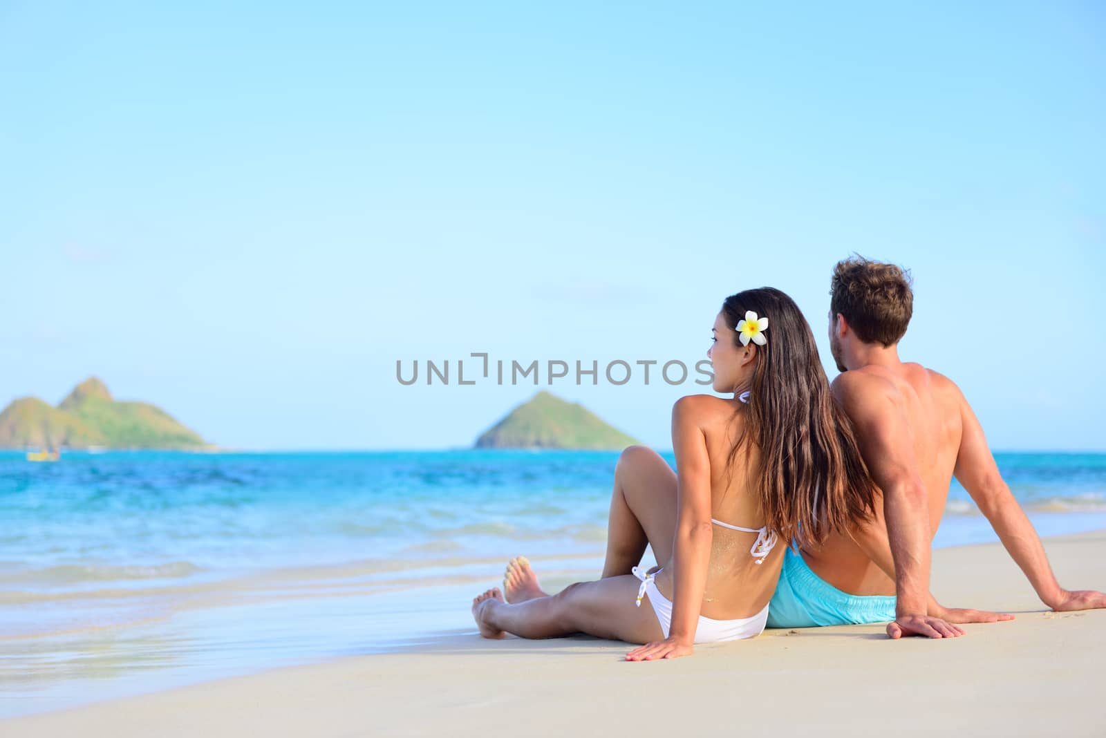 Hawaii vacation couple relaxing tanning on beach. Beautiful young adults on holidays lying down on white sand of Lanikai beach, Oahu island, Hawaii, USA.