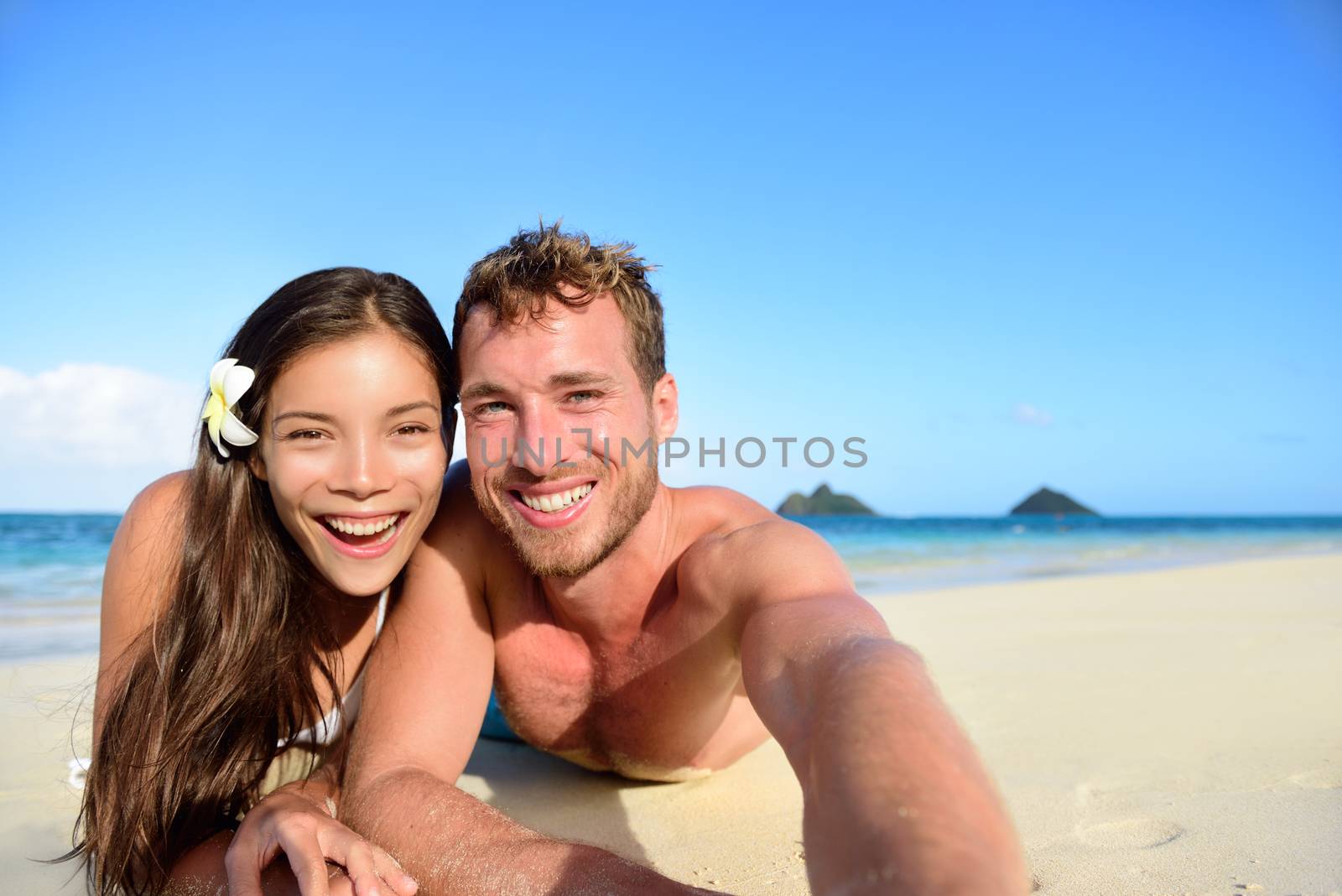 Couple relaxing on beach taking selfie picture with camera smartphone. Young multiracial couple on getaway vacation in Hawaii lying down looking at camera. Candid closeup angle looking real.