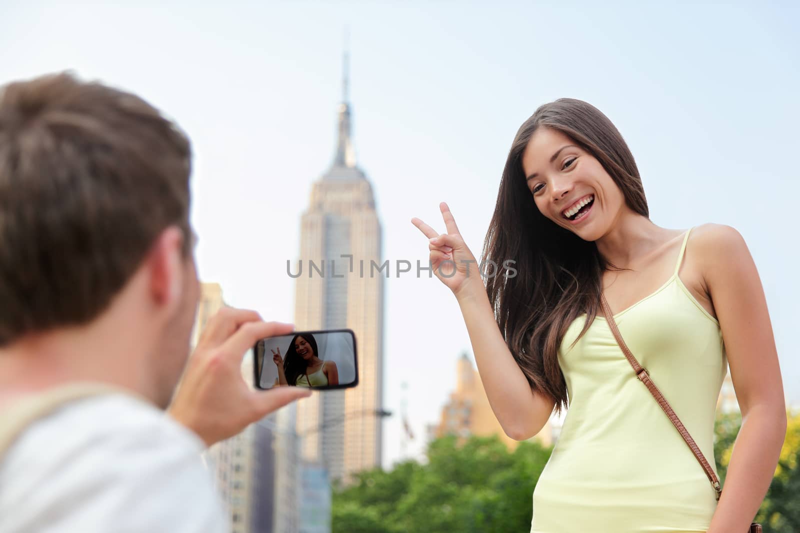 NYC asian chinese tourist girl posing at Empire State Building doing the v hand sign. Young couple of tourists taking pictures with smartphone in New York City in front of famous landmark building.