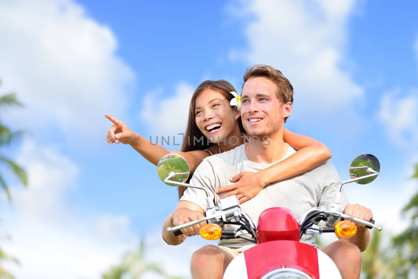 Happy couple pointing on scooter travel vacation. Young Asian woman showing her boyfriend a location while driving a moped on a road trip during summer holidays with blue sky and clouds background.
