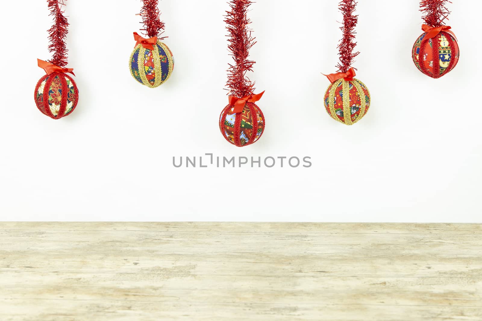Christmas copy space: five Christmas baubles made by hand with the decoupage technique hung from top with a red shiny garland on white background and light wooden base on bottom by robbyfontanesi