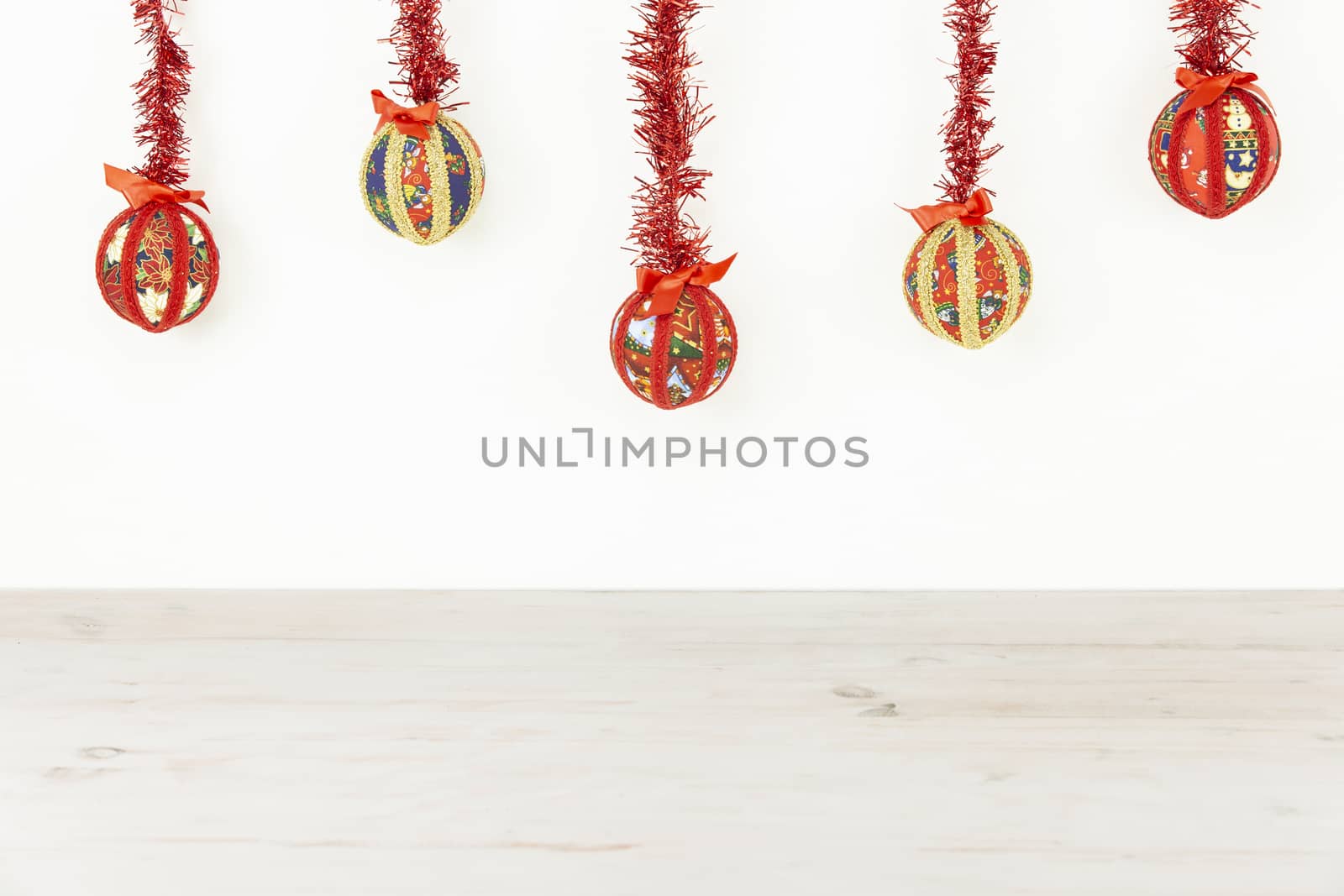 Christmas copy space: five Christmas baubles made by hand with the decoupage technique hung from top with a red shiny garland on white background and light wooden base on bottom