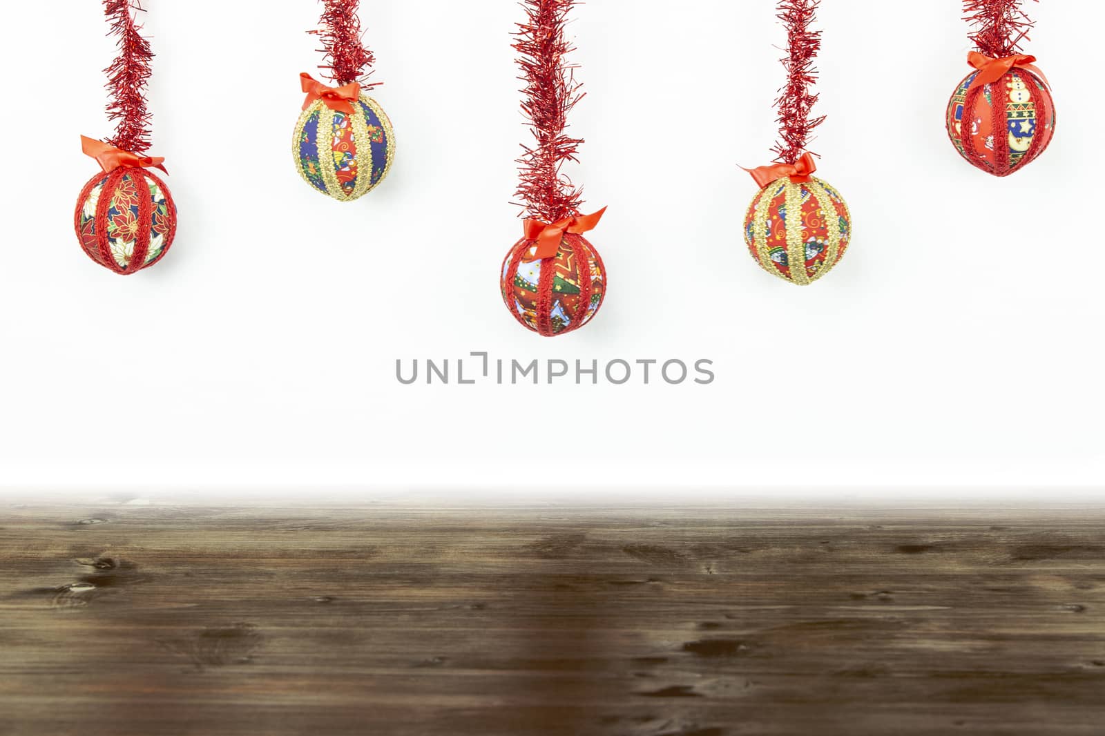 Christmas copy space: five Christmas baubles made by hand with the decoupage technique hung from top with a red shiny garland on white background and dark wooden base on bottom by robbyfontanesi