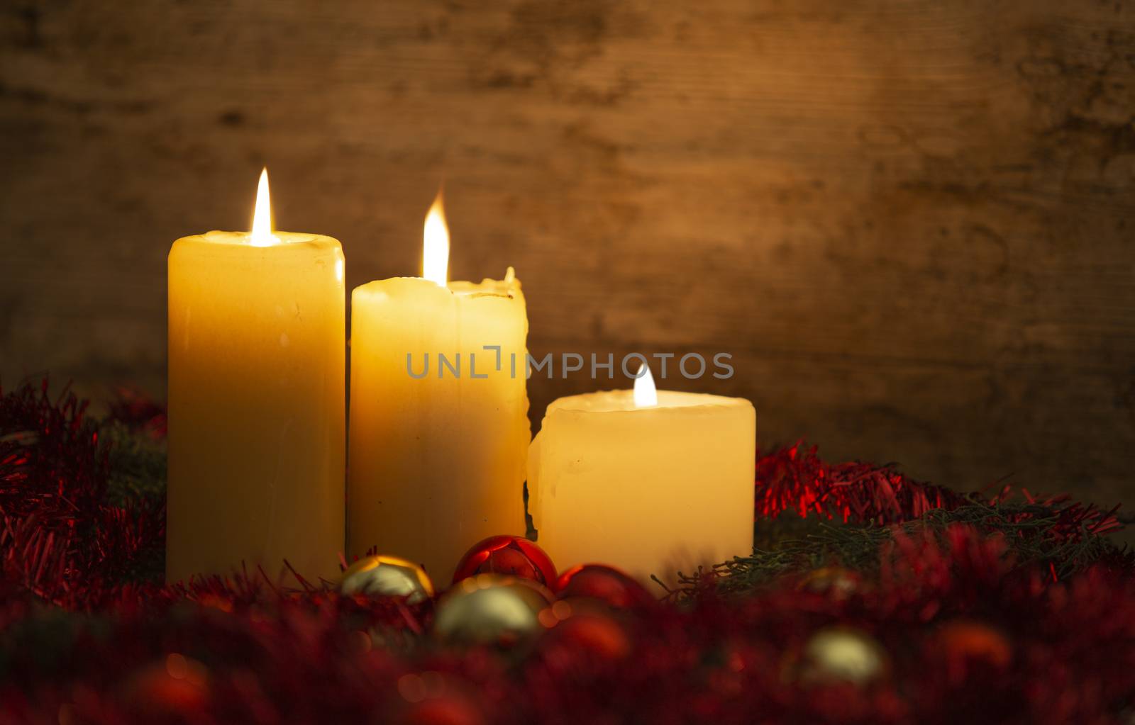 The warmth of the Christmas concept: three candles lit on a light wooden table and a rustic setting with pine branches, red decoration and gold and red bright baubles with bokeh effect by robbyfontanesi