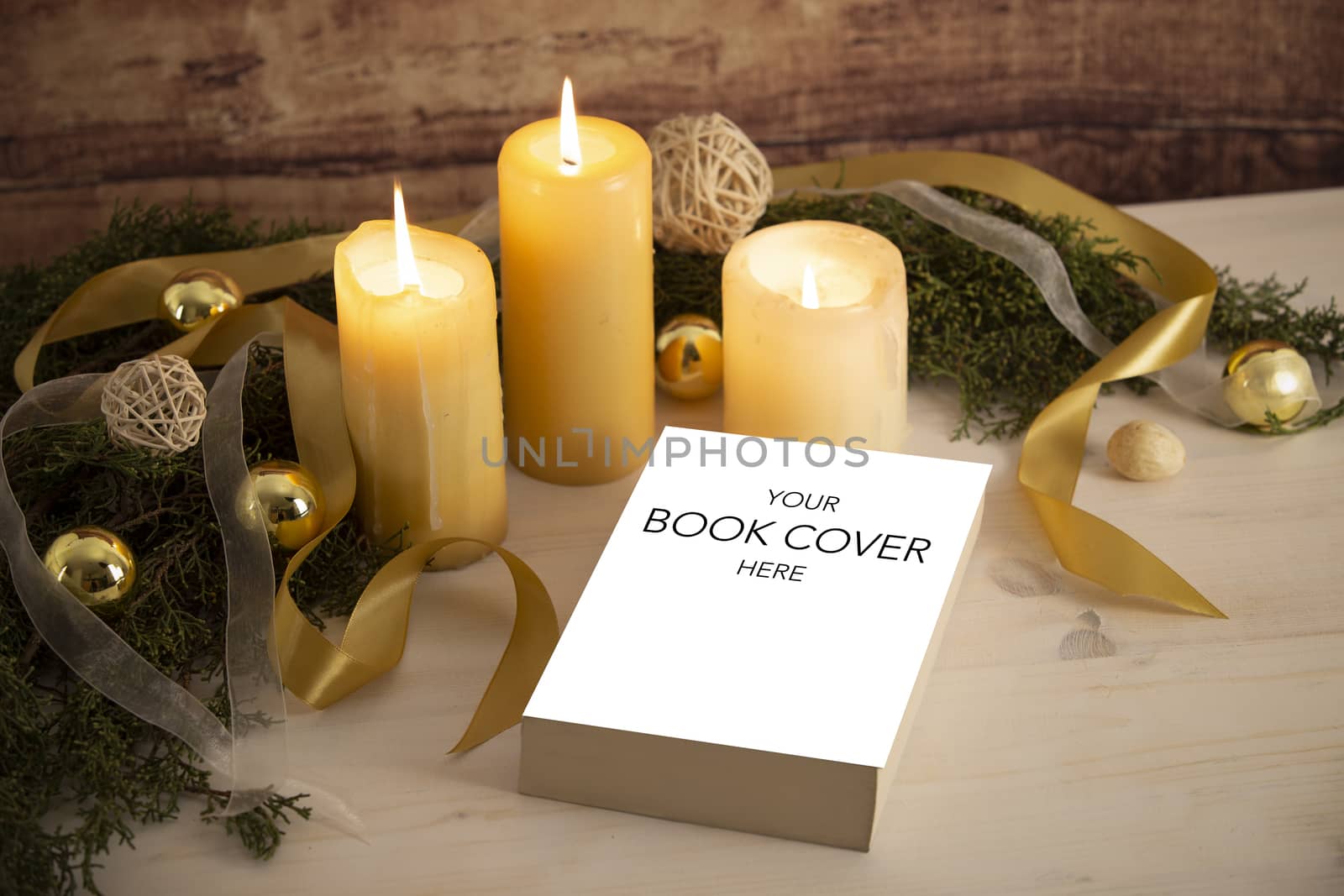 Christmas setting for a book presentation: book with blank cover settled with three lit candles , white organza and gold satin ribbons, gold baubles on light wooden table and dark wood background by robbyfontanesi