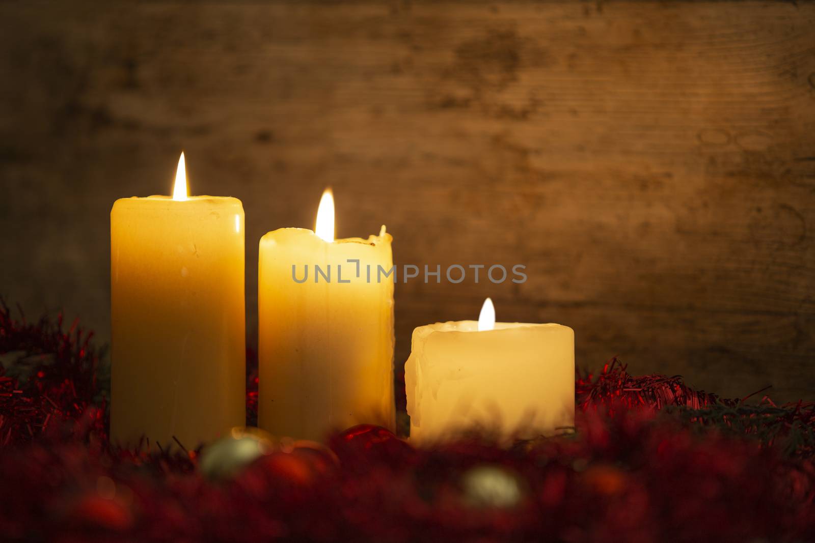 The warmth of the Christmas concept: three candles lit on a light wooden table and a rustic setting with pine branches, red decoration and gold and red bright baubles with bokeh effect