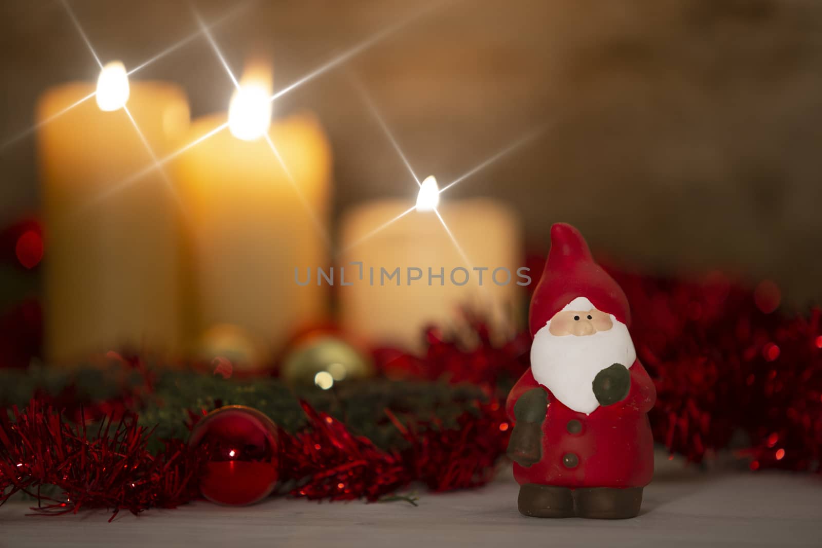 The warmth of the Christmas concept: a Santa Claus statuette in foreground in a rustic setting with with three candles lit in bokeh effect, gold and red bright baubles, cross screen star effect