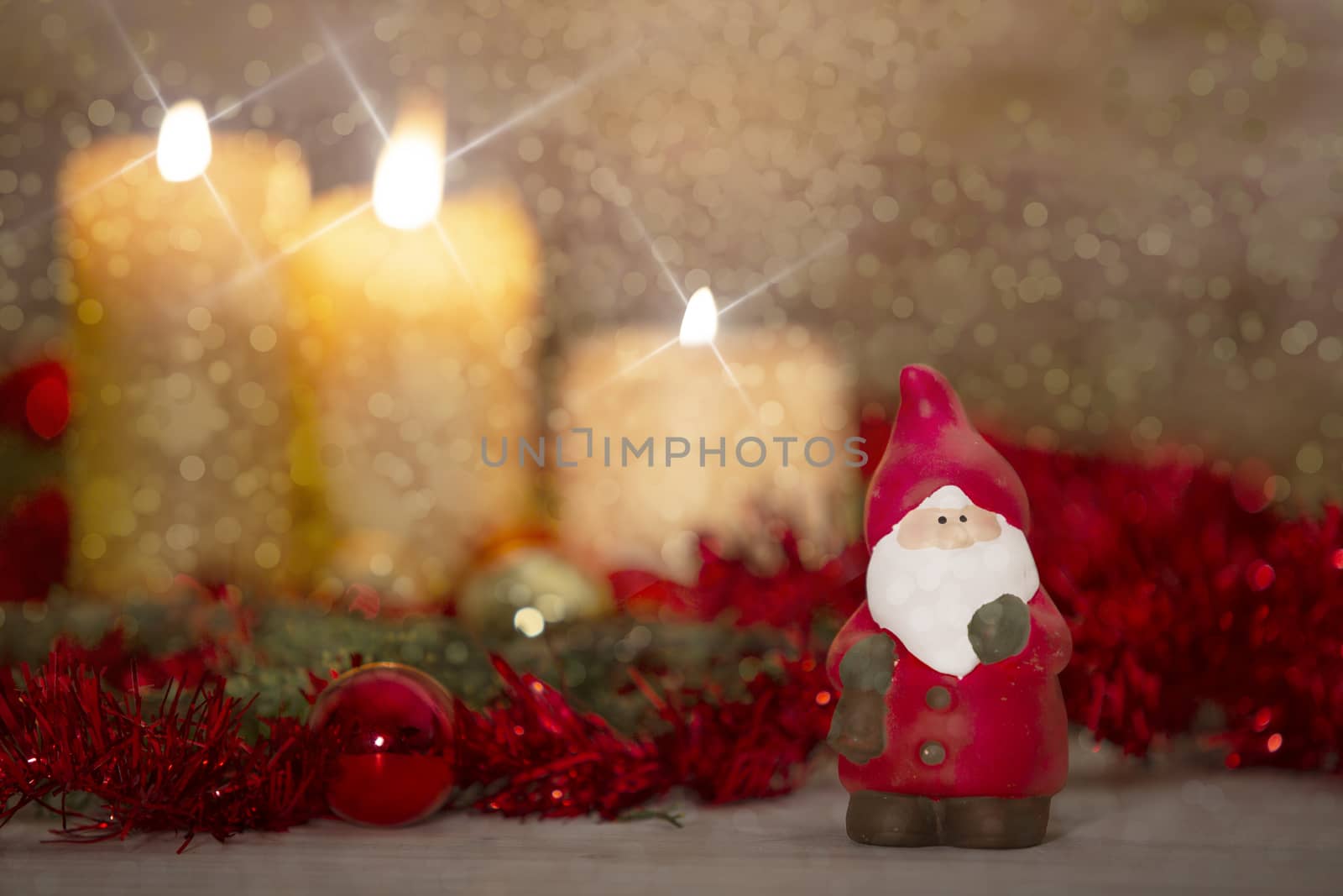 The warmth of the Christmas concept: a Santa Claus statuette in foreground in a rustic setting with with three candles lit in bokeh effect, gold and red bright baubles, cross screen and snow effect