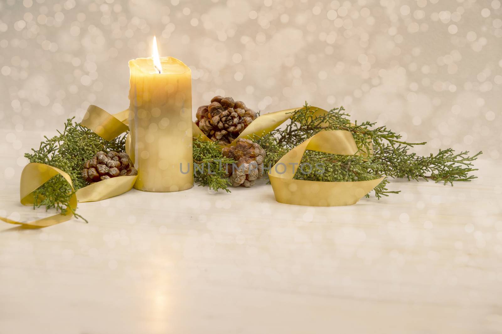 Christmas copy space with a lit yellow candle, pine branches, golden satin ribbon and natural pine cones on a light patterned background