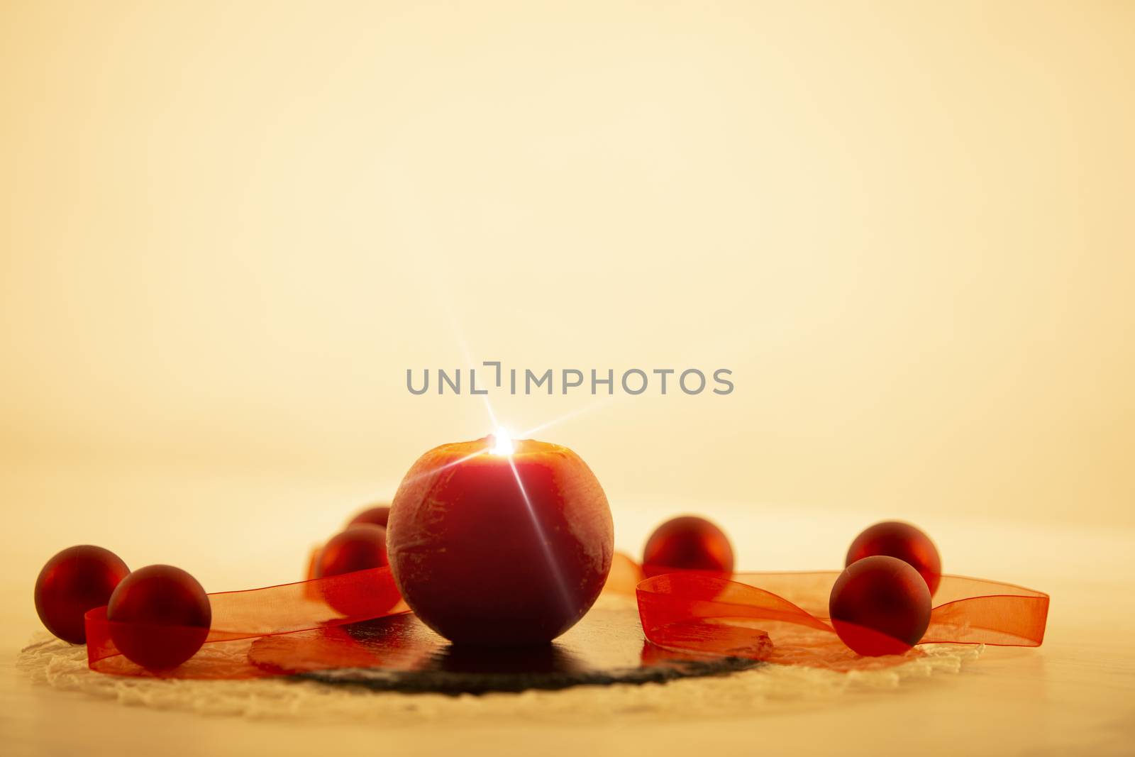 Modern Christmas copy space: close up of a red lit candle with cross screen effect on a limestone plate surrounded by red baubles and a red organza ribbon