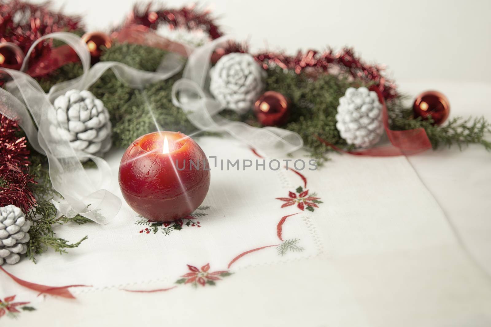 Christmas setting: a red lit candle with cross screen effect on foreground surrounded by pine branches, red baubles, red and white ribbons, white pine cones on Christmas tablecloth in bokeh effect