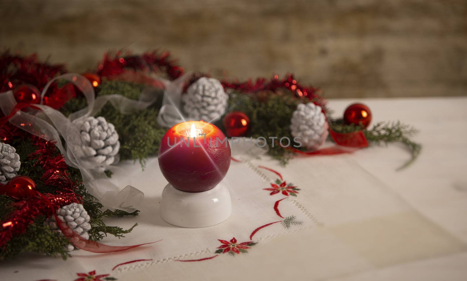 Christmas setting: a red lit candle with cross screen effect on foreground surrounded by pine branches, red baubles, red and white ribbons, white pine cones on Christmas tablecloth in bokeh effect by robbyfontanesi