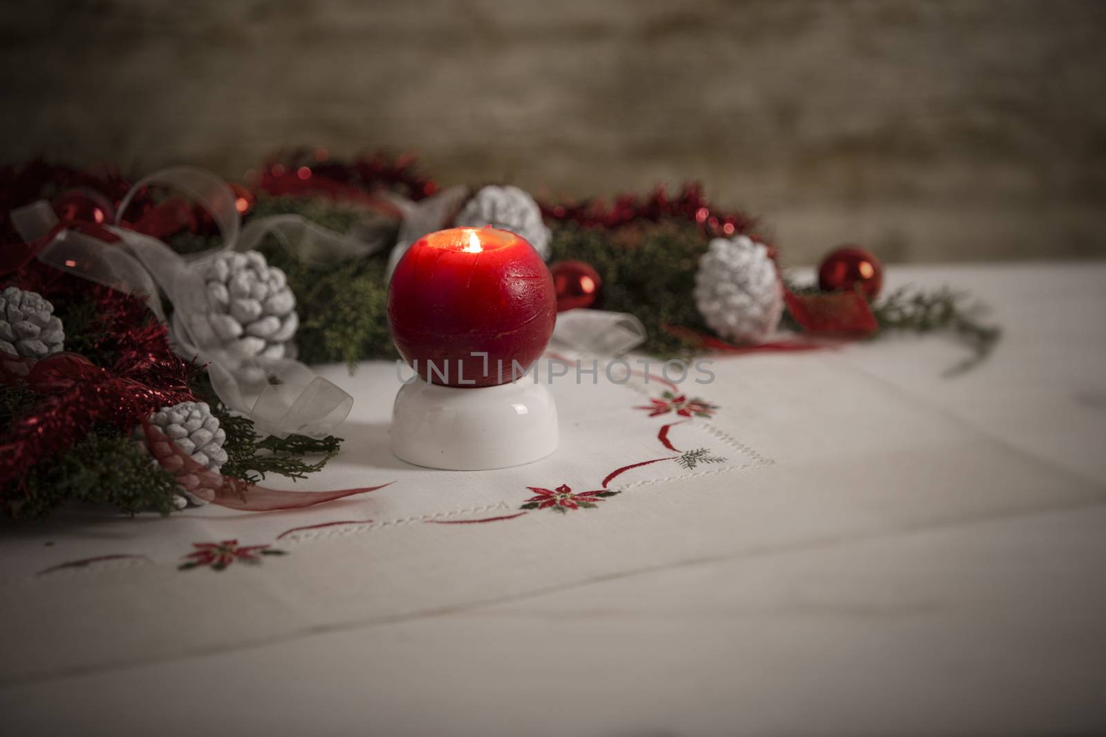 Christmas setting: a red lit candle on foreground surrounded by pine branches, red baubles, red and white ribbons, white pine cones on Christmas tablecloth in bokeh effect by robbyfontanesi