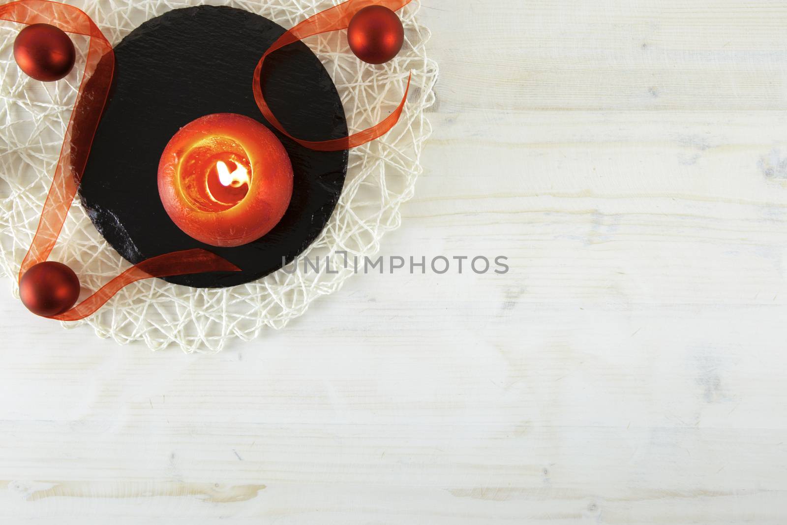 Flat lay Christmas copy space with a red organza ribbon, a red lit candle, a black round limestone plate on light wooden background by robbyfontanesi