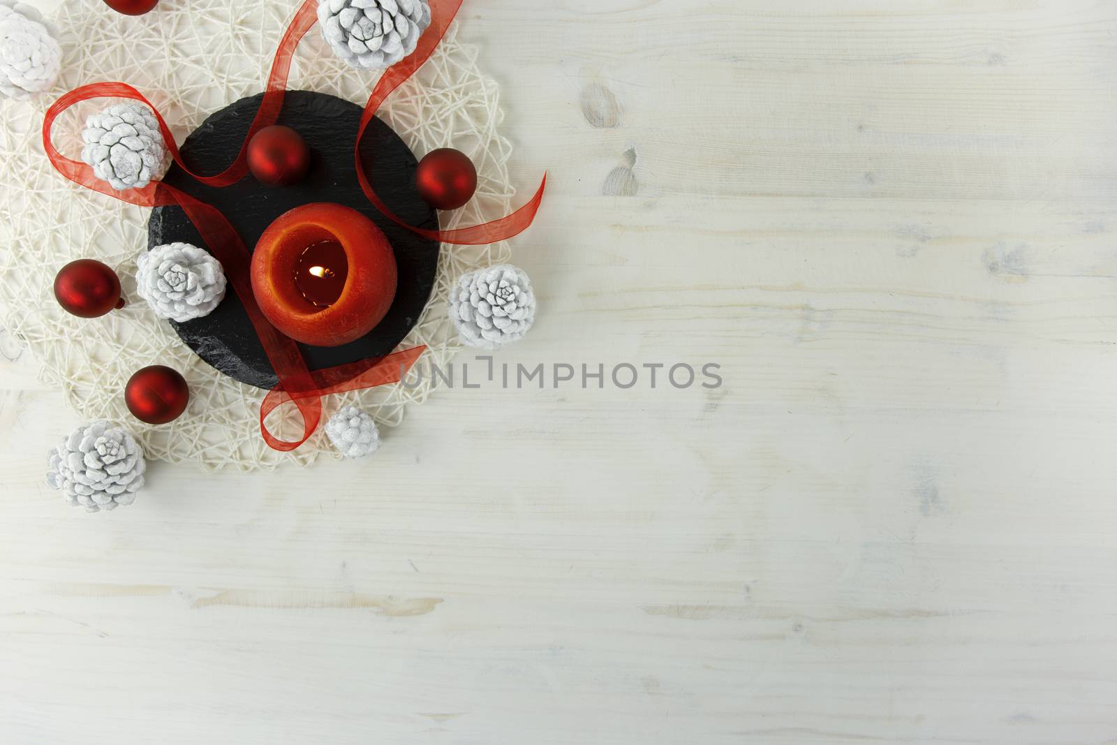 Flat lay Christmas copy space with a red organza ribbon, a red lit candle, a black round limestone plate, white pine cones on light wooden background