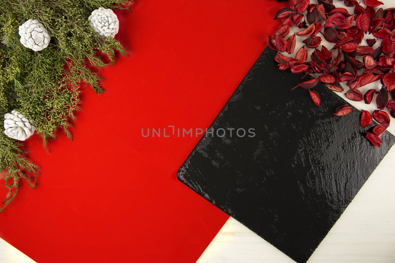 Flat lay geometric Christmas copy space with a diagonal red stripe, a black limestone plate, pine branches, white pine cones and red petals on light wooden background