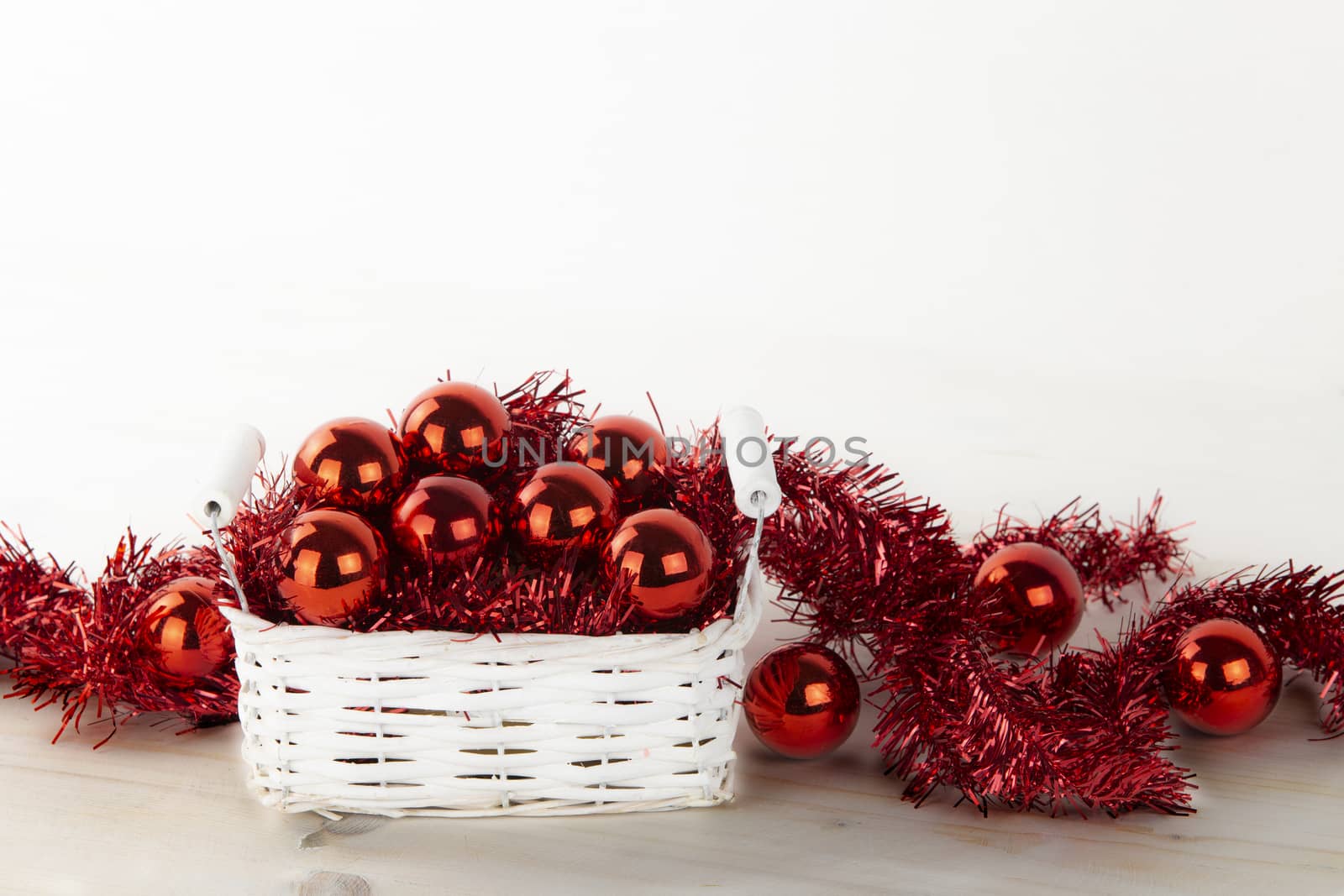 Low angle Christmas copy space: little shabby white basket full of red Christmas baubles with red decorative wreath on light wooden table and white background