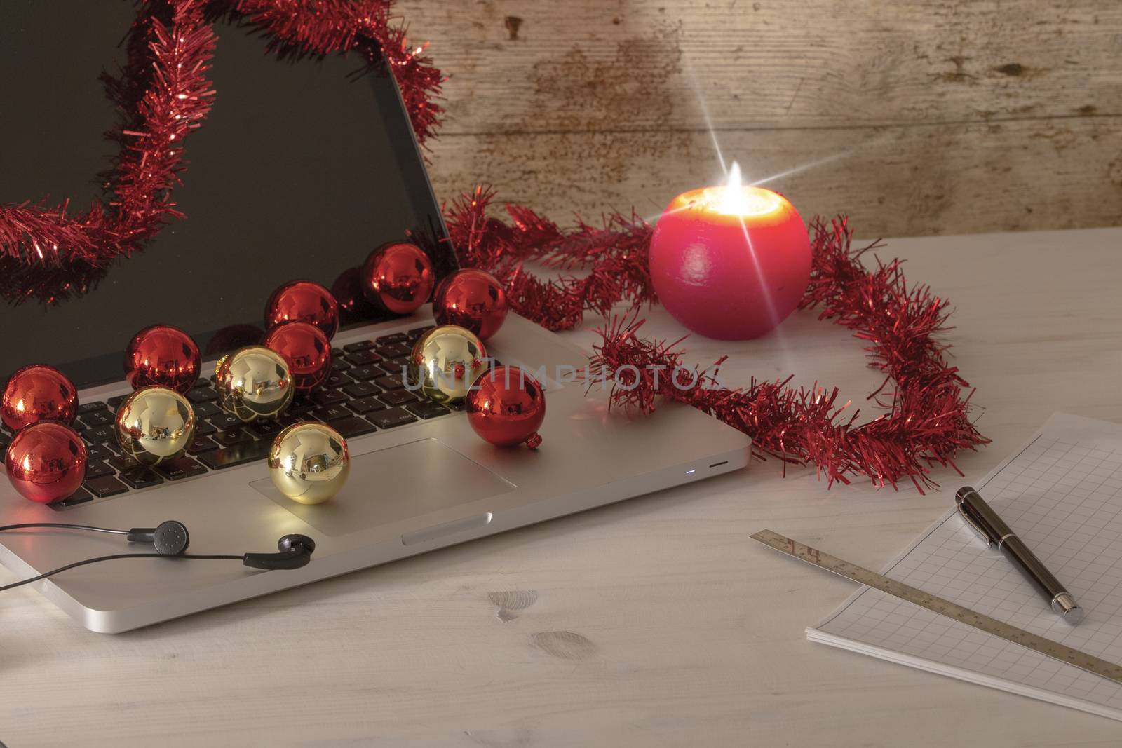 Computer job at Christmas holidays concept: an aluminum laptop open, red wreath decoration, red and gold baubles, lit candle, pen and ruler on block notes on light wooden table by robbyfontanesi