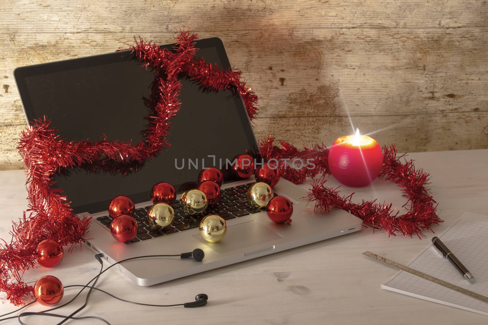 Computer job at Christmas holidays concept: an aluminum laptop open, red wreath decoration, red and gold baubles, lit candle, pen and ruler on block notes on light wooden table