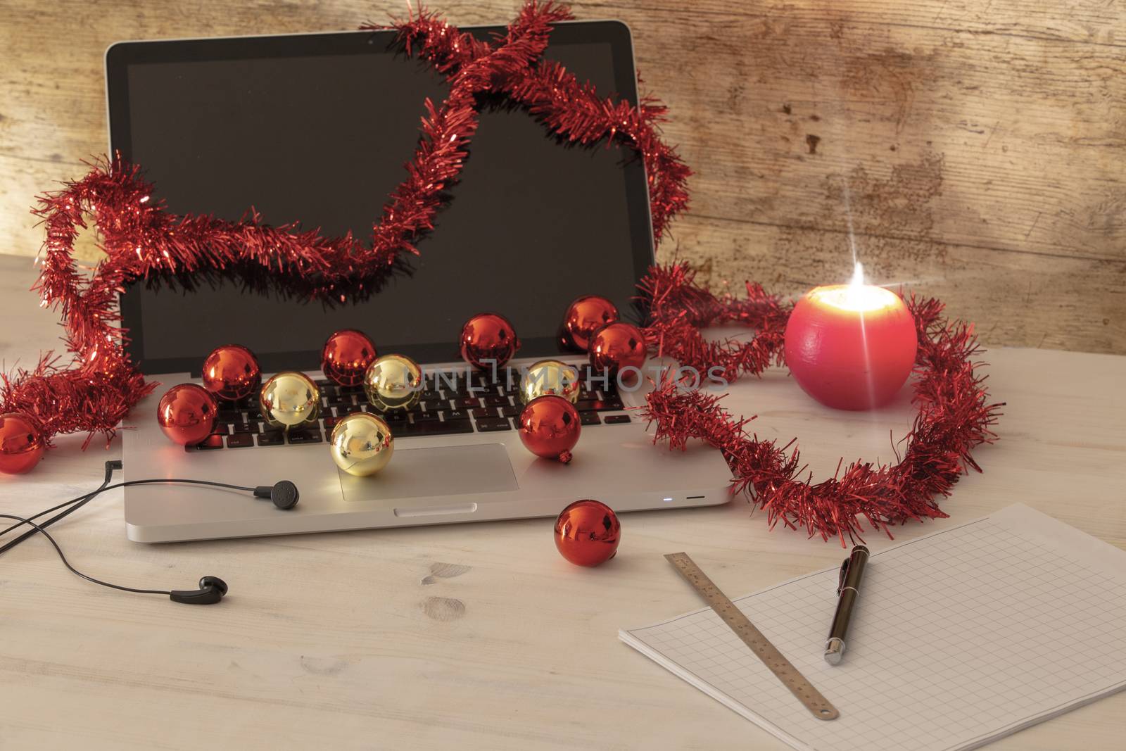 Computer job at Christmas holidays concept: an aluminum laptop open, red wreath decoration, red and gold baubles, lit candle, pen and ruler on block notes on light wooden table