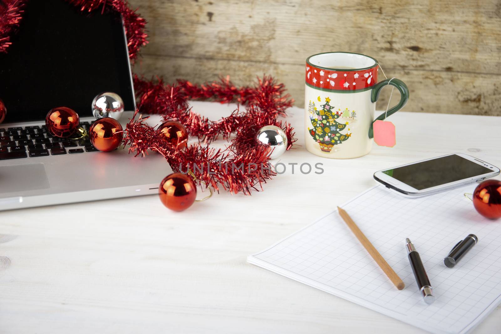 Computer job at Christmas holidays concept: light wooden table with an open aluminum laptop, red decoration, red baubles, Christmas cup of tea, smartphone, block notes, pencil and pen on block note