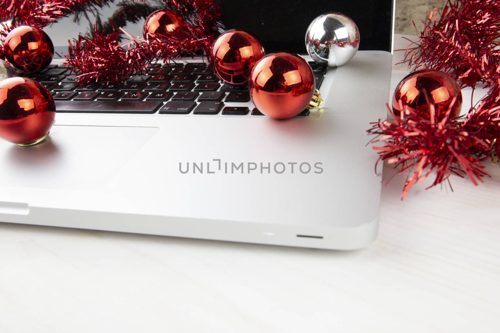 Computer job at Christmas holidays concept: close up in low angle view of an aluminum laptop open, red wreath decoration and red and silver baubles on a light wooden table