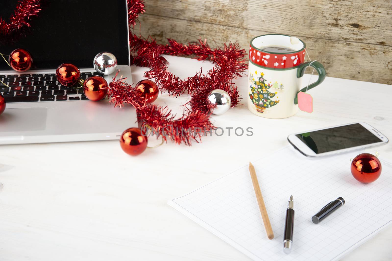 Computer job at Christmas holidays concept: light wooden table with an open aluminum laptop, red decoration, red baubles, Christmas cup of tea, smartphone, block notes, pencil and pen on block note