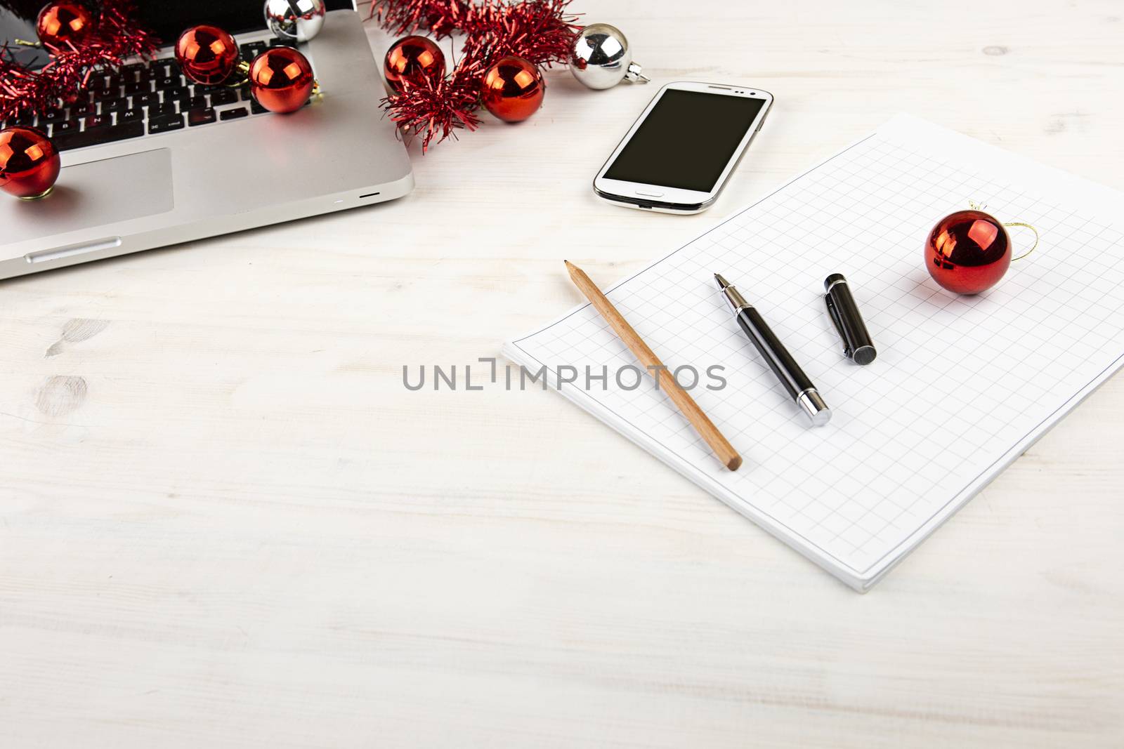 Computer job at Christmas holidays concept: light wooden table with an open aluminum laptop, red decoration, red baubles, smartphone, block notes, pencil and pen on block note by robbyfontanesi