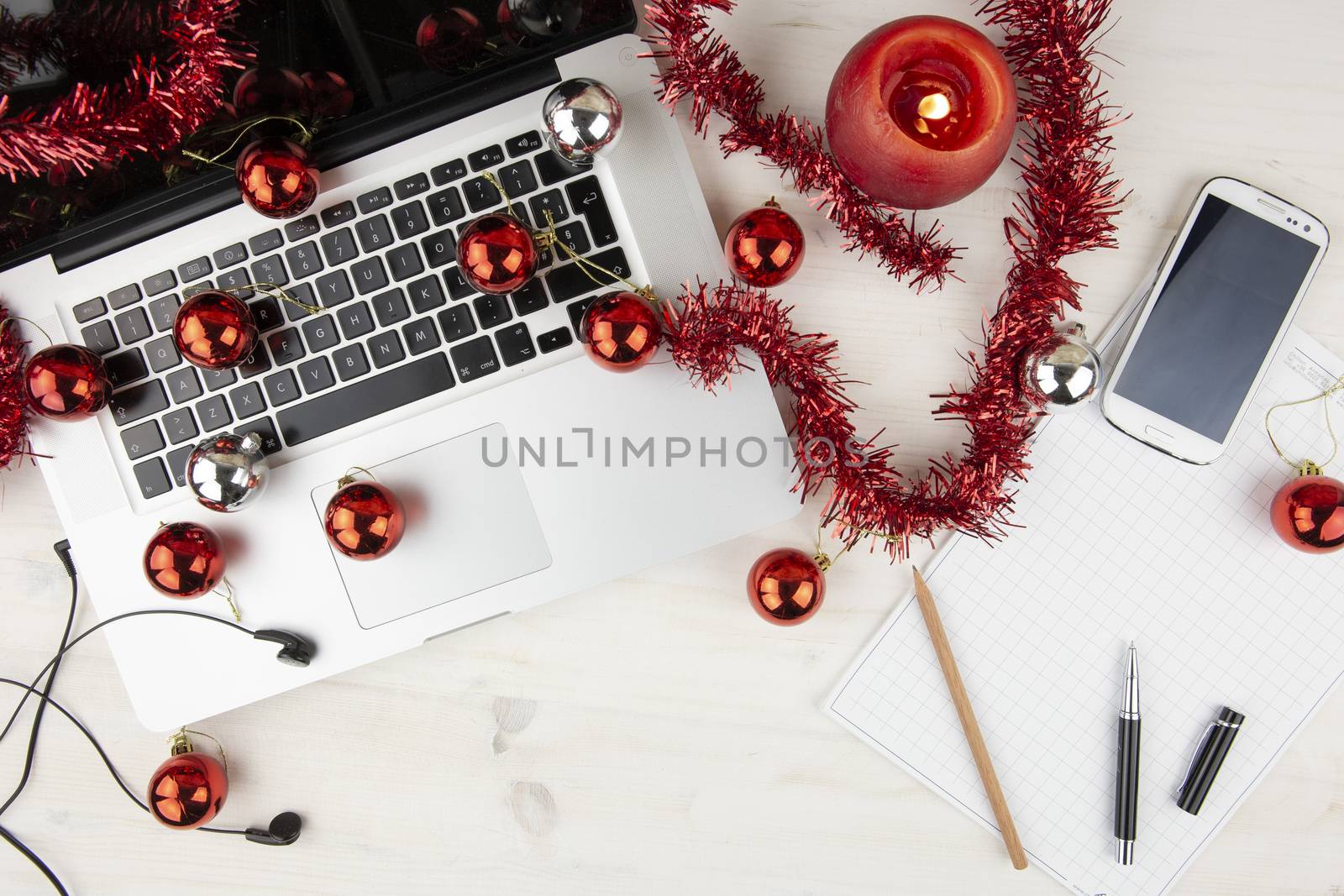 Flat lay computer job at Christmas holidays: top view of a light wooden table with an aluminum laptop open, red decoration, red baubles, a smartphone, lit candle, block notes, ear pods, pencil and pen by robbyfontanesi