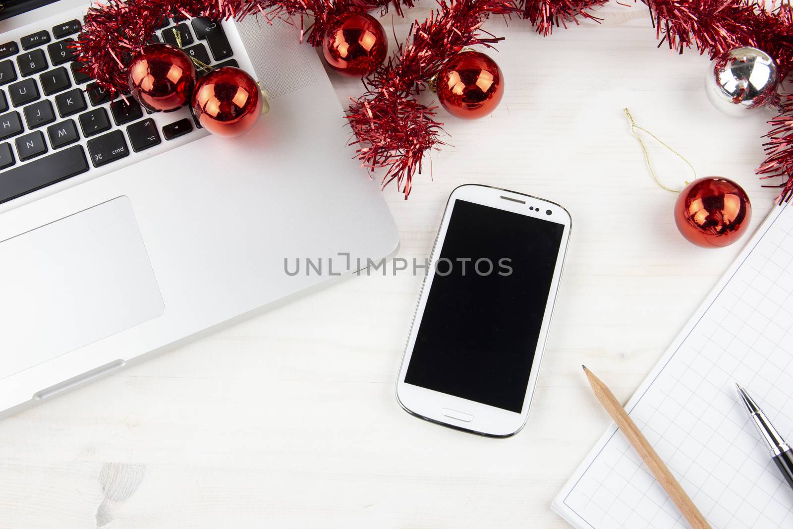 Computer job at Christmas holidays concept: close up top view of an aluminum laptop open, smartphone, red wreath decoration, red and silver baubles, pen and pencil on block notes on light wooden table by robbyfontanesi