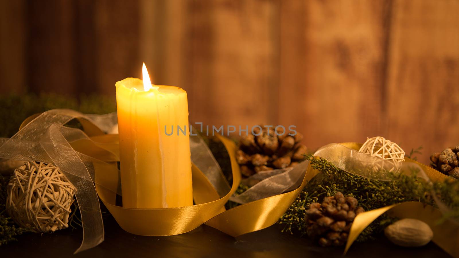 The warmth of the Christmas concept: one candle lit on a dark wooden table and a rustic wooden setting with pine branches, natural pine cones and gold satin and white organza ribbons