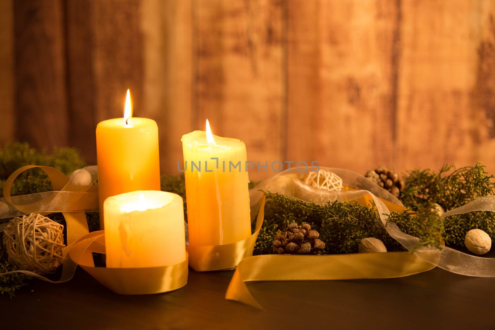 The warmth of the Christmas concept: three candles lit on a dark wooden table and a rustic wooden setting with pine branches, natural pine cones and gold satin and white organza ribbons