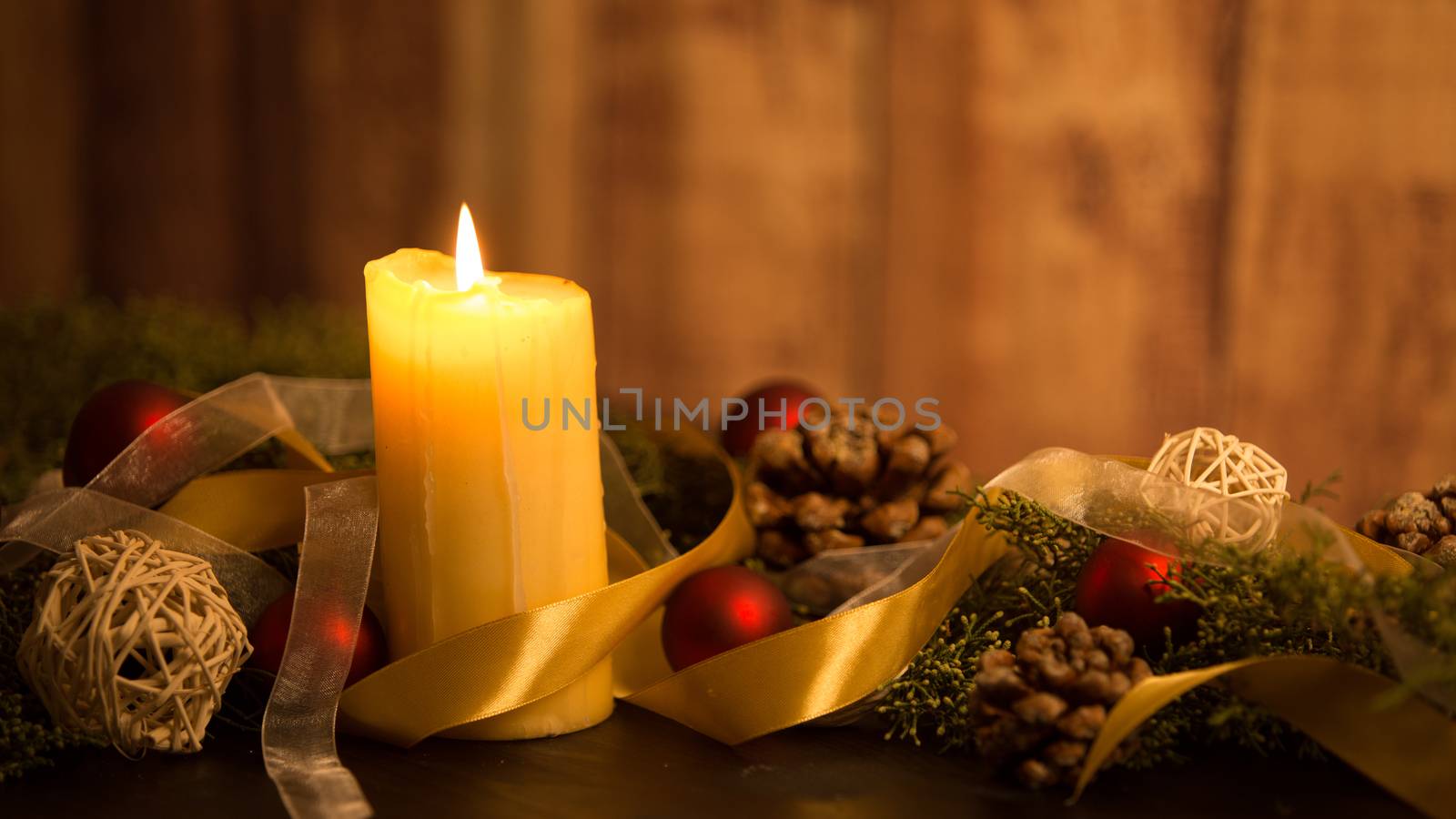 The warmth of the Christmas concept: close up of three candles lit on a dark wooden table and with pine branches, natural pine cones and gold and red bright baubles with a gold satin ribbon by robbyfontanesi